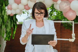 Mulher de óculos e blazer branco utiliza um tablet enquanto organiza um evento decorado com balões - Organização de Eventos Blog Campus